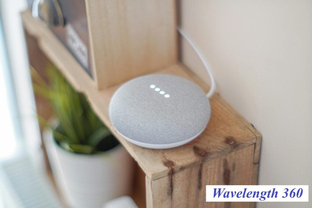 A close-up shot of a small, grey fabric-covered smart speaker with its indicator lights on, sitting on a wooden shelf, representing a common Internet of Things (IoT) device that requires a compact, high-performance custom antenna.